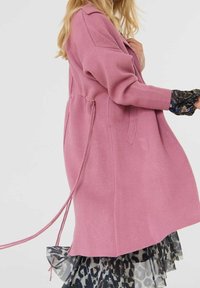 Femme en manteau rose ceinturé à manches longues, portant une jupe à volants imprimé animal et des poignets fleuris, vue de côté sur un fond uni.