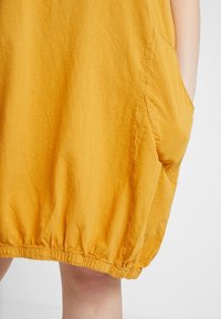 Gul, lätt klänning med en rynkad fåll och sidofickor. Tyget har en mjuk, texturerad yta. Elastik vid fållen ger struktur.