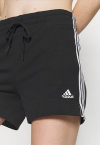 Pantalones cortos deportivos negros hechos de tela suave, con franjas blancas a los lados y un logo bordado de Adidas en el dobladillo inferior izquierdo.