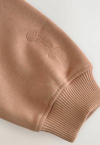 Poignet de sweatshirt rose clair avec texture côtelée et logo brodé "Sports Club" en fil légèrement plus foncé. Tissu doux, semblable à du jersey.