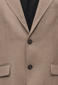 Καφέ υφασμάτινο σακάκι με δύο μαύρα κουμπιά, πέτο με σχισμή και τσέπες με καπάκι πάνω από μαύρο πουκάμισο.