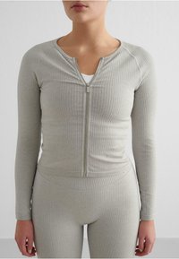 Siva rebrasta zip-up majica s dugim rukavima, koja ima okrugli izrez i uski kroj. Materijal izgleda mekano sa suptilnom teksturom.