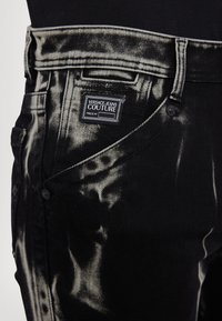 Melni džinsi ar izbalējušu, nolietotu rakstu. Iezīmēti ar Versace Jeans Couture etiķeti un metāla pogu akcentiem.