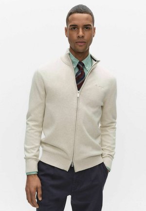 Ljusbeige zip-up tröja i mjukt tyg med hög krage och ribbade muddar, bärs över en grönrandig skjorta och slips.