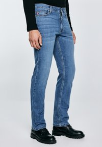 Mees, kes kannab siniseid sirgete sääreosadega pükse ja musti nahast paeltega kingi, ühevärvilise tausta ees.