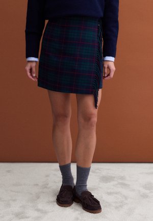 Una modelo lleva una falda cruzada de cuadros en verde oscuro y azul marino con flecos, un jersey azul marino, calcetines grises y mocasines marrones con borlas.