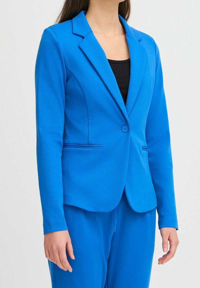 Femme portant un blazer bleu vif cintré avec un seul bouton et un pantalon bleu assorti par-dessus un top noir, debout devant un fond uni.