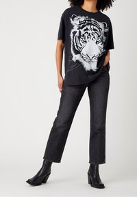 Henkilö, joka pukeutuu mustaan T-paitaan, jossa on suuri valkoisen tiikerin kasvokuva, mustiin farkkuihin ja mustiin korkean varren saappaisiin, seisoo valkoista taustaa vasten.