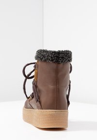 Bota de cuero marrón con una suela texturizada en color beige, cuello de forro polar gris y cordones oscuros. Presenta detalles de ante en los laterales.