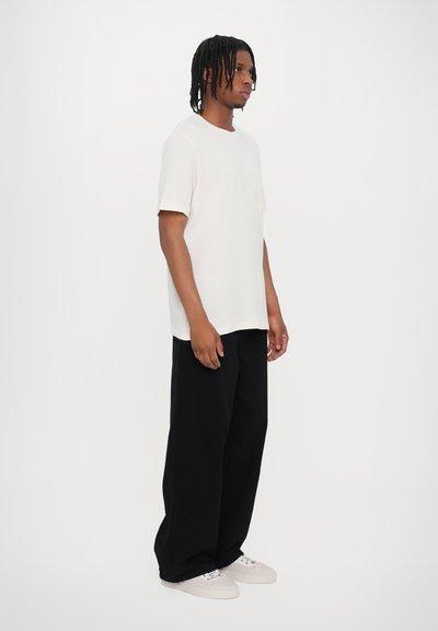 Vit kortärmad t-shirt med rund halsringning, parat med löst sittande svarta byxor och vita sneakers, med en avslappnad passform och minimalistisk design.