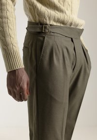 Pantalones de corte ajustado en color verde oliva con una cintura alta, pinzas y un cinturón de tela con hebilla dorada. Suéter de punto texturizado en color crema visible.