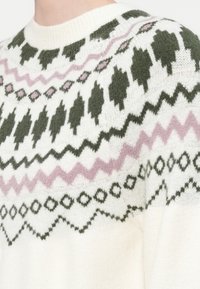 Bela pletena jopica z zelenimi in roza geometričnimi vzorci po prsih in ramenih, ki ima okroglo ribasto ovratnico.