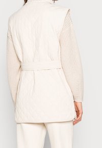 Gilet matelassé beige clair avec une taille ceinturée, présentant une texture lisse et un motif en losanges, associé à des manches et un ourlet tricotés.