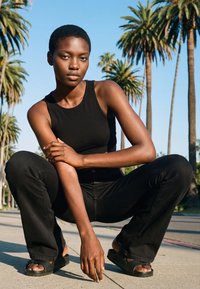 Tânăr îmbrăcat în maiou negru și pantaloni, aplecat în poziție ghemuită pe trotuar, cu palmieri și cer albastru în fundal.