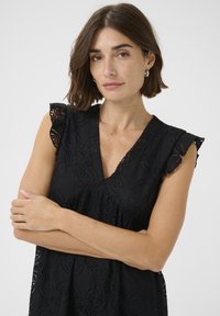 Rochie neagră cu detalii din dantelă florală, decolteu în V și mâneci cu volane evazate. Textură de material moale, design strâns care accentuează silueta.