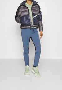 Granatowa kurtka puchowa o textured wykończeniu, jasnozielona koszulka, niebieskie spodnie z czarnymi akcentami oraz jasnozielone sneakersy.