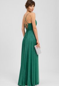 Vrouw in een groen glitterjurk tot op de vloer met kruislingse bandjes aan de achterkant, die een kleine zilveren clutch vasthoudt, poserend tegen een neutrale achtergrond.