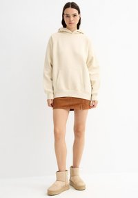 Sudadera con capucha color crema y bolsillo delantero, combinada con una falda marrón texturizada y botas cortas color canela. Ajuste sencillo y relajado.