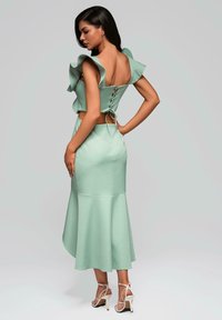 Robe vert menthe avec des manches à volants, dos lacé et ourlet évasé. Portée avec des talons blancs à lanières sur un fond neutre.