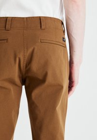 Brune chinos med en glat tekstur, der har en flad talje, bæltestropper og to baglommer. Subtil branding på baglommen.