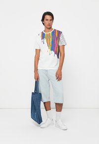 Ung mann som har på seg en hvit T-skjorte med fargerike abstrakte mønstre, lyse denimshorts, hvite joggesko, og holder en blå denim totebag.