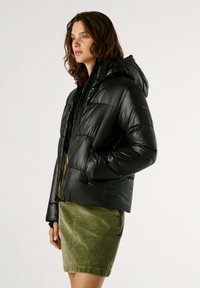 Zwarte pufferjas met capuchon, met een glanzende textuur en gestikte ontwerp. Gecombineerd met een groene corduroy rok.