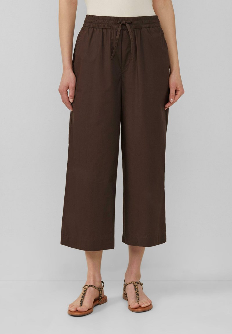 Bruine, wijde broek met een elastische tailleband en een trekkoord. Gemaakt van lichte stof, met zijzakken. Gepaard met bruine sandalen.