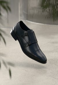 Sort læder double monk strap dress sko med sølv spænder, præsenteret over en tekstureret lys grå overflade og sløret baggrund.