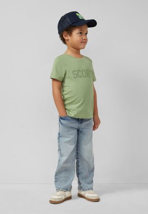 Młody chłopiec w zielonej koszulce z napisem "Score", jasnoniebieskich dżinsach, białych trampkach i granatowej czapce, stojący z rękami w kieszeniach, spoglądający na bok.