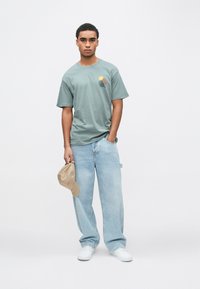 Gaiši zaļa kokvilnas T-krekls ar saules un viļņu grafiku, kopā ar gaiši ziliem brīva piegriezuma džinsiem un rokā turētu bēša krāsas beisbola cepuri.