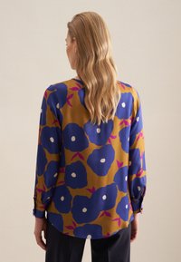 Camicia floreale di colore giallo senape con grandi fiori blu e bianchi, con maniche lunghe e dettagli con bottoni sui polsini. Tessuto morbido.