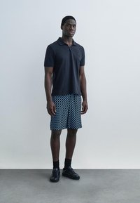 Moški stoji, oblečen v črno polo majico, modre kratke hlače z vzorcem, črne nogavice in črne superge, na enobarvnem ozadju.