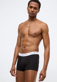 Boxer shorts in cotone nero con una cintura bianca che presenta il logo "TOMMY HILFIGER". Il modello ha un fisico definito e un tono di pelle naturale.