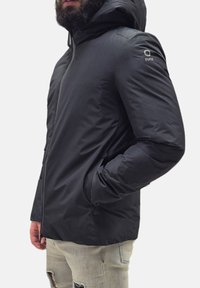 Uomo con barba che indossa un giubbotto invernale con cappuccio nero e jeans strappati grigi chiari, in piedi con una mano nella tasca del giubbotto.