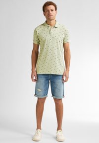 Světle zelené polo tričko s malými černými vzory, zkombinované s ohrnutými džíny s efektem opotřebovaného vzhledu. Celkový look doplňují bílé tenisky.