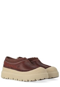 Coppia di scarpe marroni slip-on con suole spesse beige, punte testurizzate e logo UGG sul lato, inclinate verso destra.