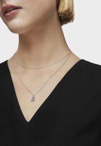 Collana in argento a strati con una delicata catena e un pendente testurizzato, indossata sopra un top nero a scollo a V. Lo sfondo è bianco uniforme.
