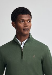 Grön zip-up tröja i mjukt material, med krage, ribbade ärmslut och en liten broderad logotyp på bröstet.