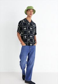 Camisa negra de manga corta con un patrón de hojas de palma blancas, combinada con jeans azules holgados y un sombrero de cubo verde. Unos zapatos negros completan el conjunto.
