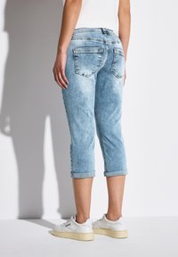 Un jean capri en denim bleu clair avec un ourlet retroussé et des poches arrière ; porté avec des baskets blanches. Le tissu a un motif texturé et délavé.