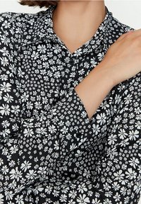 Schwarze Kurzarmbluse mit floralen Mustern in Weiß, mit einem Kragen und langen Ärmeln, die verschiedene Blumenmuster präsentieren.