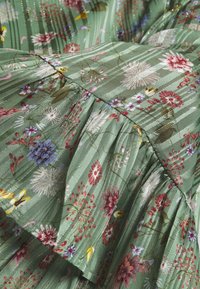 Tessuto con stampa floreale verde e design plissettato, caratterizzato da fiori di vari colori su uno sfondo a strisce. Accenti scintillanti visibili.