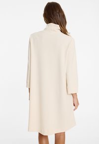 Eine Frau trägt einen cremfarbenen, knielangen Mantel mit hohem Kragen und Dreiviertelärmeln, von hinten zu sehen.