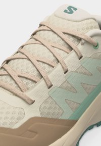 Κοντινό πλάνο σε μπεζ και ανοιχτό πράσινο αθλητικό παπούτσι με υφή υφάσματος, κορδόνια στο ίδιο χρώμα, έντονο λογότυπο στο πλάι και ενισχυμένη σόλα.