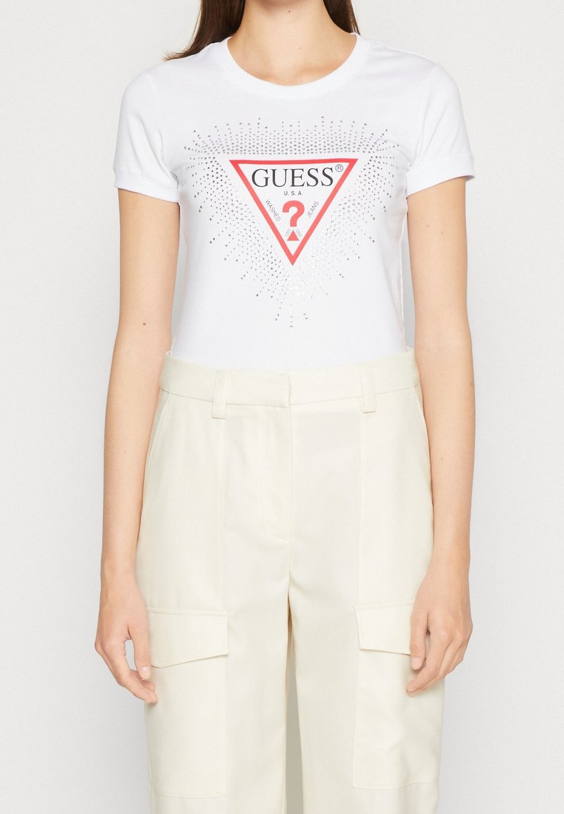 Femme portant un t-shirt blanc Guess avec un logo triangle rouge et un pantalon beige taille haute avec poches latérales à rabat.