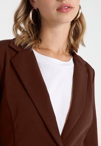 Hněd�ý blazer s klopou, nosený přes bílou tričko s kulatým výstřihem. Modelka má vlasy po ramena a velké kruhové náušnice.