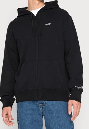 Homme portant un sweat à capuche noir zippé avec des poches avant et un petit logo blanc sur la poitrine, associé à un jean bleu.