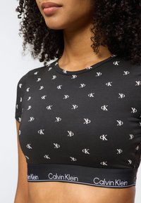 Γυναίκα που φοράει μια εφαρμοστή μαύρη κοντή μπλούζα με διασκορπισμένα λευκά λογότυπα "cK" και ελαστική ταινία με τη μάρκα Calvin Klein στο τελείωμα.