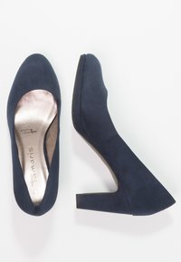 Escarpins en daim bleu marine avec un embout rond et un talon épais. L'intérieur présente une finition métallique. Texture lisse.