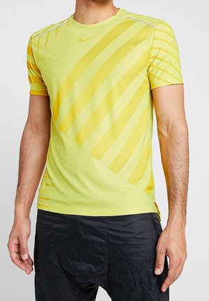 Mężczyzna ubrany w żółtą sportową koszulkę z krótkim rękawem z ukośnymi pasami oraz czarne, teksturowane szorty sportowe, stojący na tle jednolitego tła.